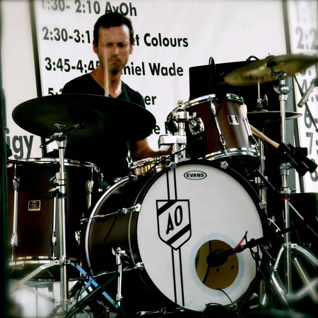 Photo of Andrew Ortolano playing the drums. He is making a face and sweating his ass off.